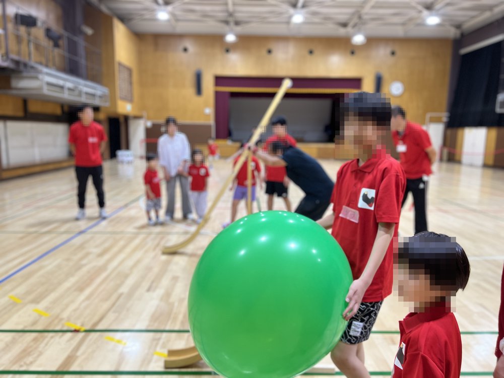 体育館で、赤いポロシャツの子どもが大きな緑のボールを持っている。うしろでは長い棒を使ってほかの子どもたちが遊んでいる