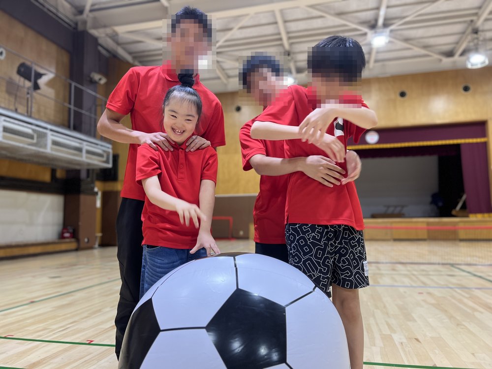 大きなサッカーボールをかこんで、Rioとサポーターのお兄さんたちが並んでいる。Rioは笑顔で両手を前に出している