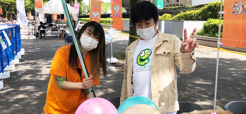 オレンジ色のTシャツを着た人と、風船を持った人がピースサインで並んでいる