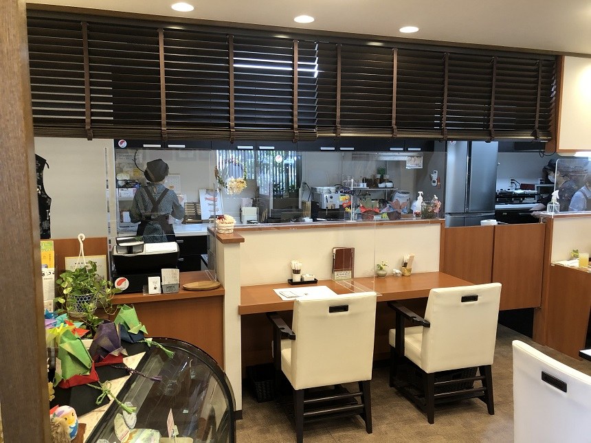 木のカウンター席と白いいすが並ぶ店内。奥にキッチンが見える