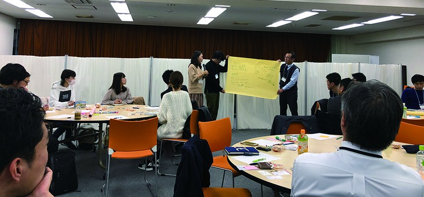 会議室の丸テーブルを囲んで話し合う参加者たち。奥で模造紙を広げている人がいる