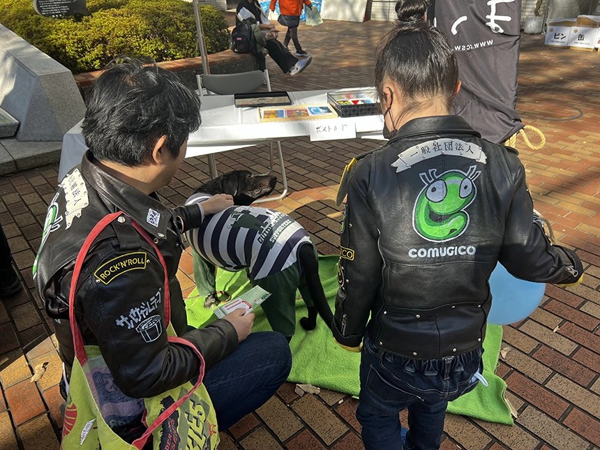 背中にCOMUGICOのロゴをペイントした革ジャンを着た親子の後ろ姿。盲導犬とふれあっている