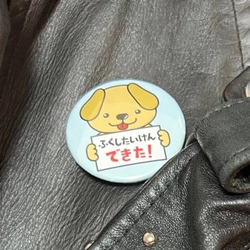 犬のイラストと「ふくしたいけん できた!」と書かれた水色の缶バッジ