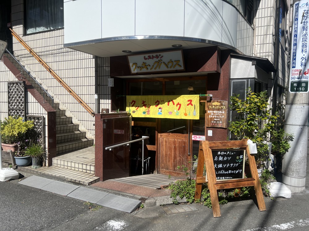ビルの1階にあるレストランの入口。黄色い手作りの暖簾に「クッキングハウス」の文字。入口の横には手すりと、黒板の「本日のメニュー」、緑の植木が並ぶ
