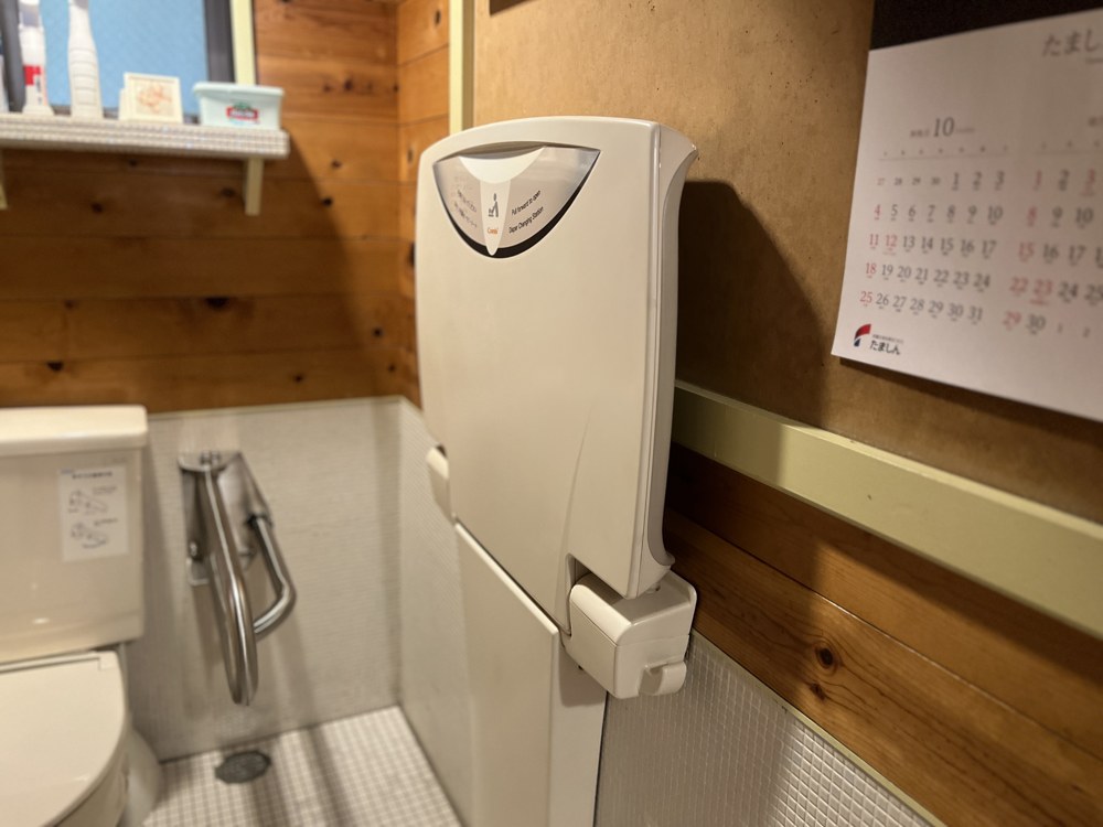 壁に折りたたまれた白い台。そばに便器と手すり、カレンダーがある