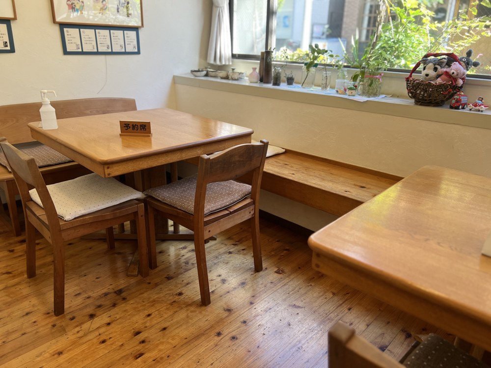 木のテーブルと椅子の席。窓ぎわにベンチがあり、窓辺には植物とぬいぐるみの入ったかごが飾られている。テーブルには「予約席」の札