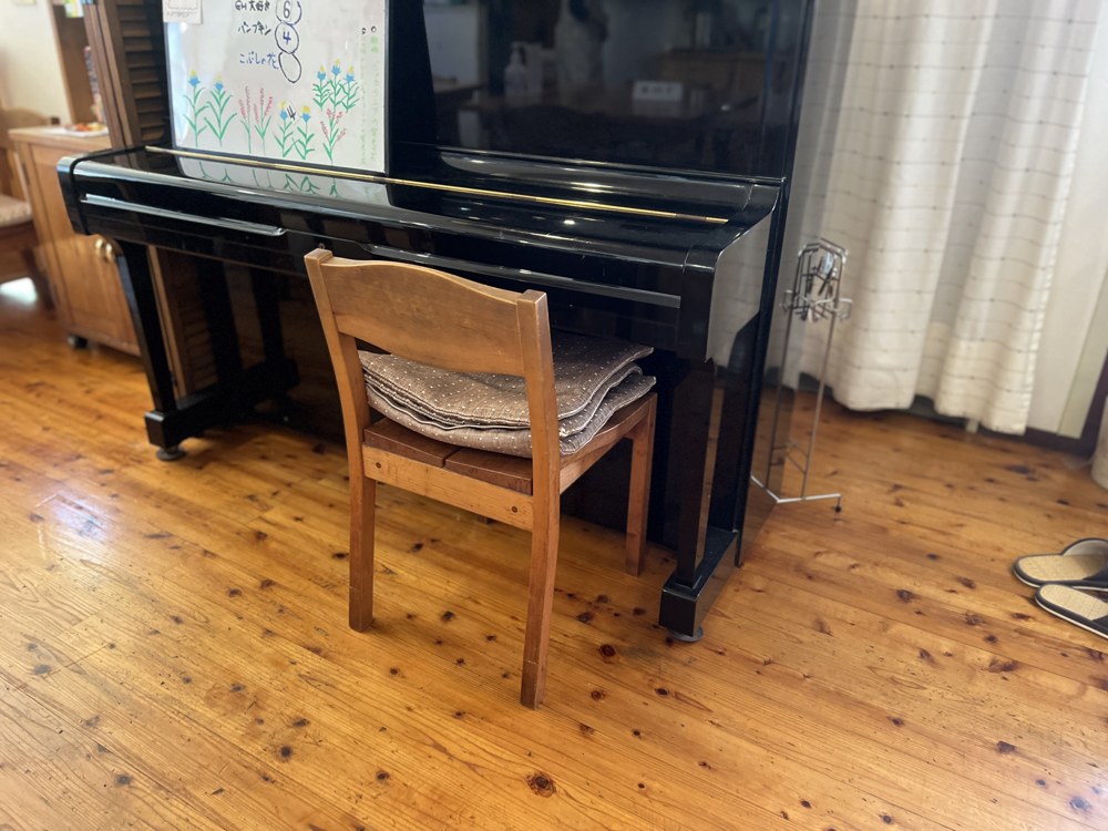 木の床の店内に置かれたピアノと、座布団のついた木の椅子。ピアノの上には手描きの花の絵が貼られている