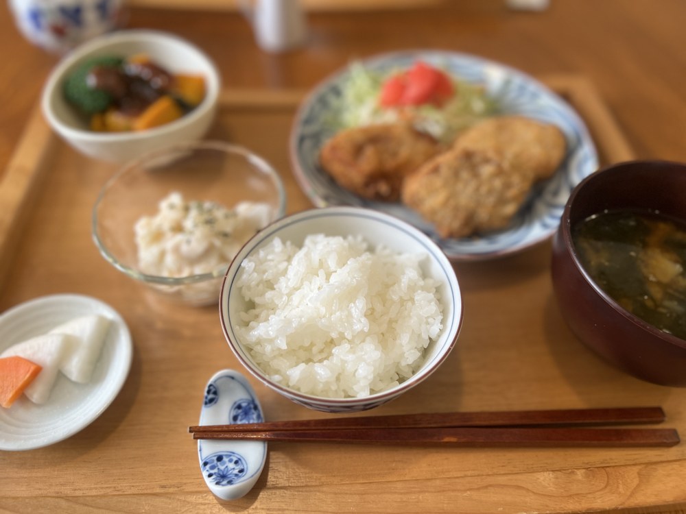 お盆に並んだランチ。白いごはん、わかめのお味噌汁、キャベツとトマトを添えた揚げ物、煮物の小鉢、大根サラダ、お漬物、お箸と青い箸置き
