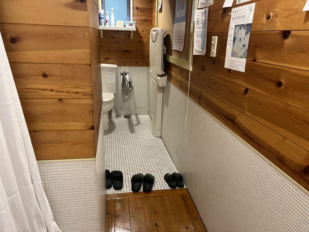 奥に長いトイレの室内。白いタイルの壁と木の床で、手前にスリッパが並んでいる。奥に便器がある