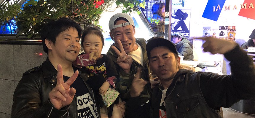 ライブハウスの前で、娘を抱いた父と男性たちがピースする記念写真