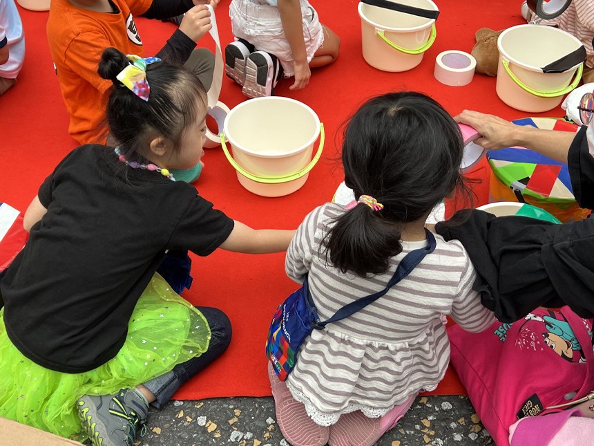 赤いシートの上で、女の子と並んでポリバケツにガムテープを貼る娘