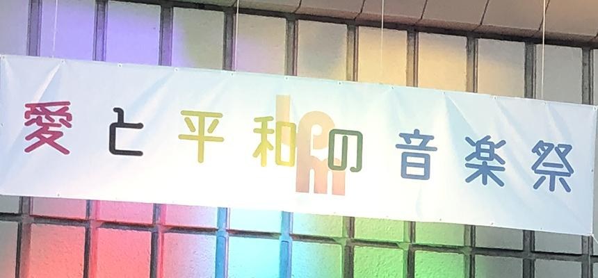 「愛と平和の音楽祭」と書かれた横断幕。後ろの窓に虹色の光