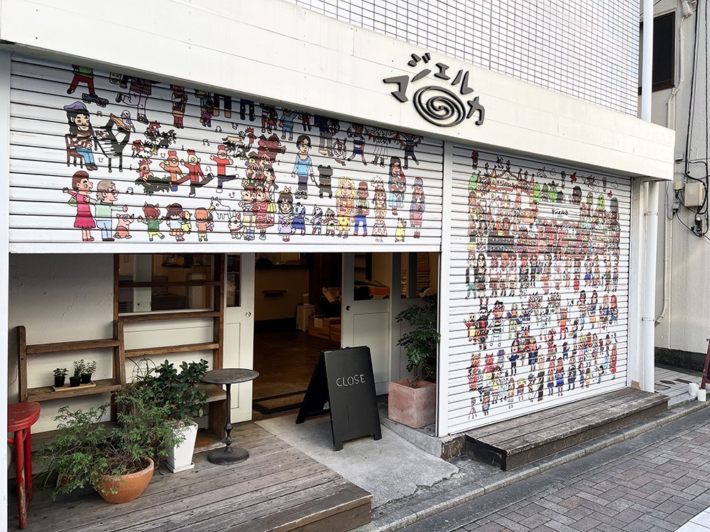 人の絵が描かれたシャッターが並ぶマジェルカの店先と、木のデッキの入口