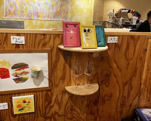 木の壁の棚に並ぶ、カラフルな額に入った顔の絵とハンバーガーの絵