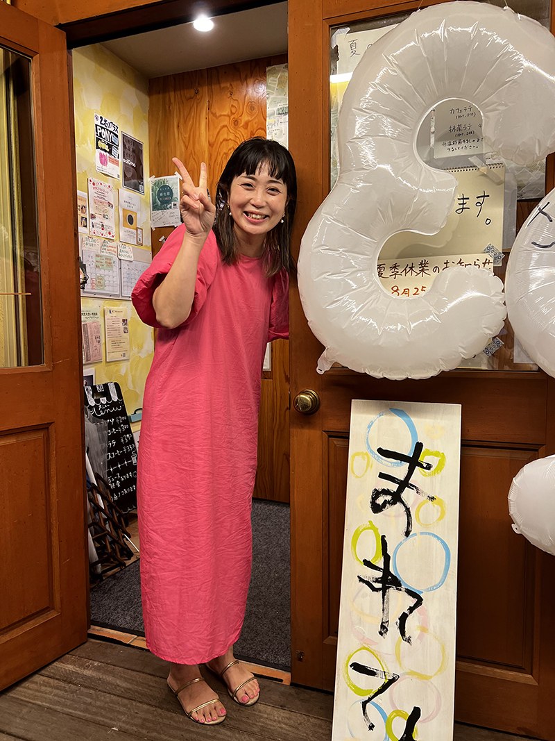 入口で風船と「まれてん」の看板のそばに立ち、ピースするマキさん