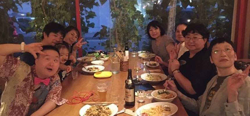 窓際の長いテーブルで料理を囲み、ピースサインで笑う仲間たちの記念写真
