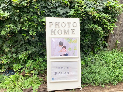 つたの生け垣の前に置かれた「PHOTO HOME」の立て看板。親子の写真が貼られている