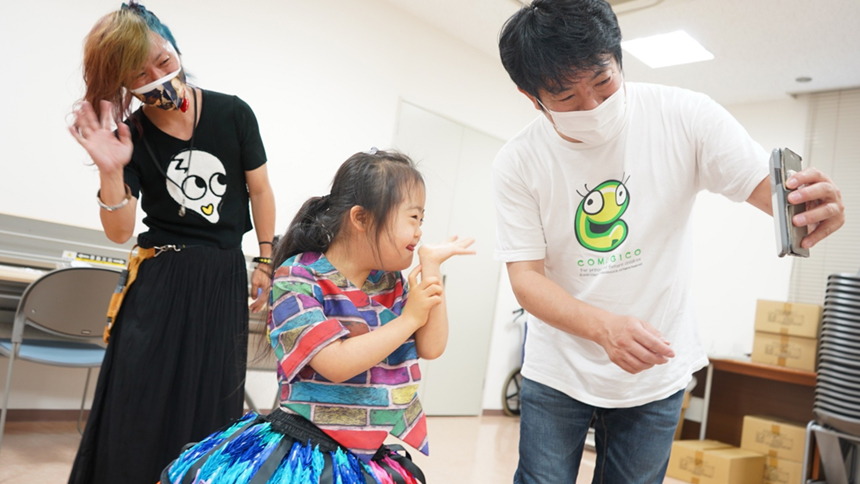 楽屋で、千羽鶴のドレスを着た子どもとスタッフたちが笑っている