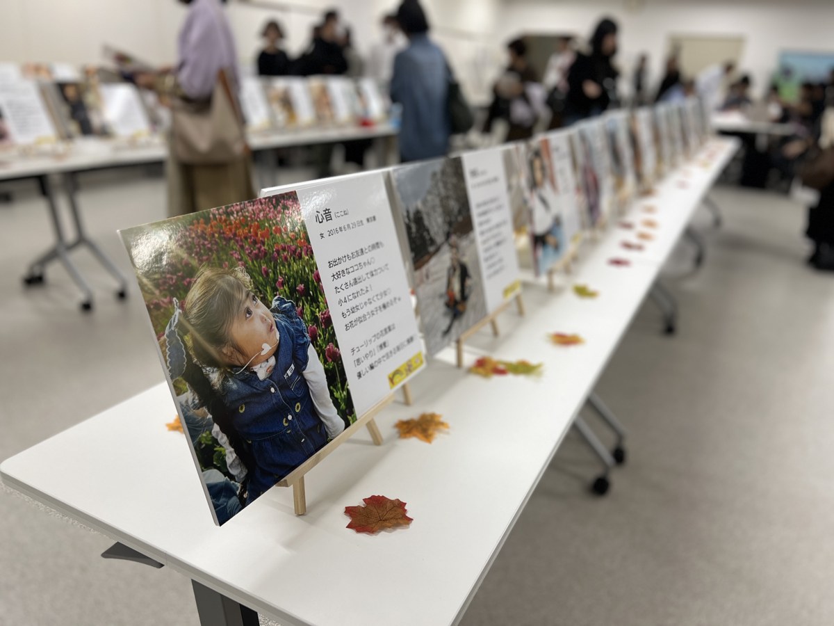 白い長机の上に、18トリソミーの子どもたちの写真と家族の言葉を添えたパネルが、イーゼルに立てられてずらりと並んでいる。あいだに秋の葉の飾り。奥では来場者がパネルを見ている