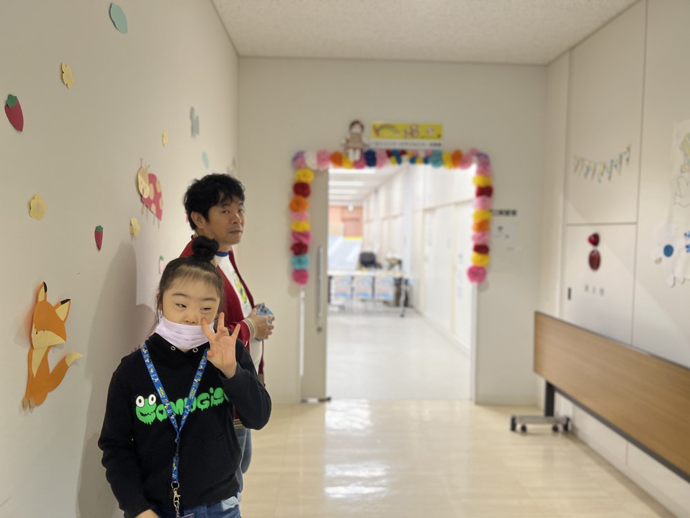 色紙の花や動物で飾られた白い廊下。奥の入口には色とりどりの花のアーチ。COMUGICOのパーカーを着たRioがピースをして、うしろにコムタクが立っている
