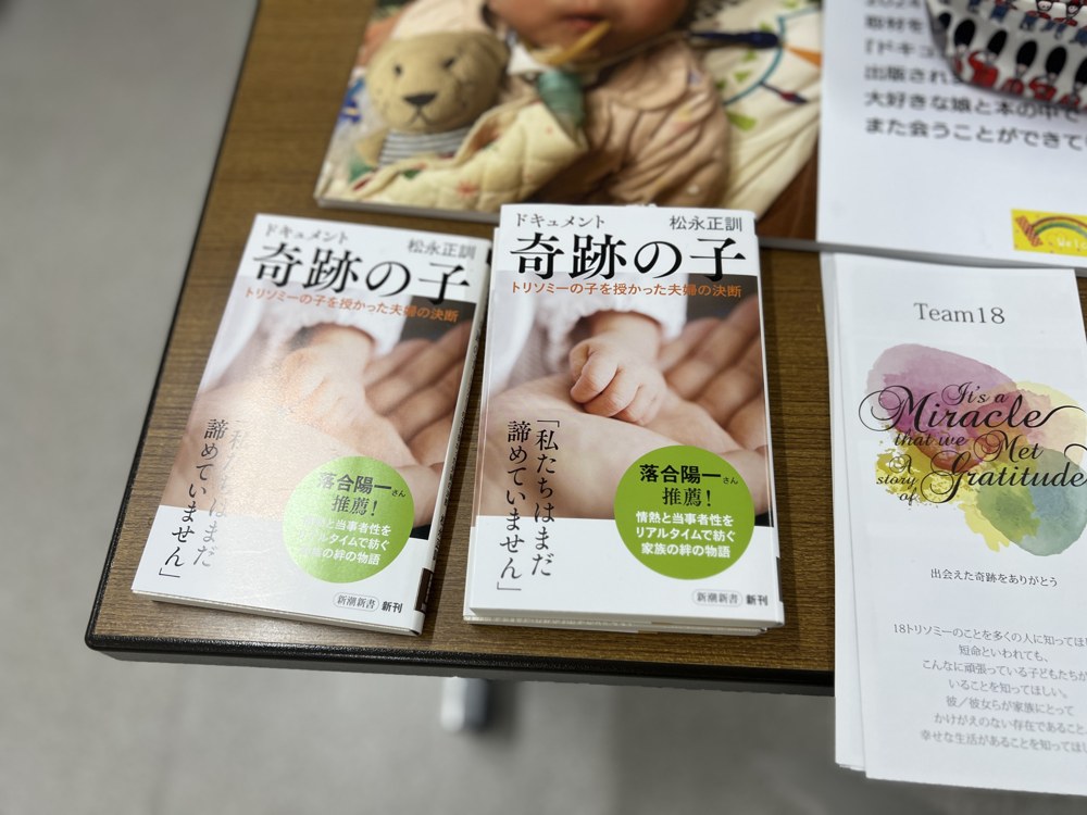 机に並んだ新潮新書『ドキュメント 奇跡の子 ―トリソミーの子を授かった夫婦の決断―』松永正訓 著。帯には「私たちはまだ諦めていません」。となりに Team18 のパンフレット