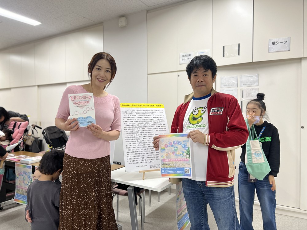 写真集『18トリソミーの子どもたち』を持った希ちゃんのお母さんの Emi さんと、写真展のチラシを持ったCOMUGICOのTシャツのコムタク。うしろでRioがピースをしている