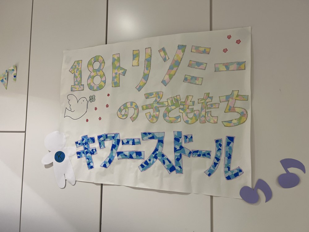 ちぎり絵で文字を作った手作りの看板。「18トリソミーの子どもたち」「キワニスドール」と書かれ、白いハトと白い布の人形が添えられている