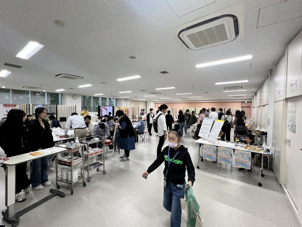 広い実習室の全景。壁ぎわの長机に写真パネルが並び、たくさんの来場者が見てまわっている。手前をCOMUGICOのパーカーを着たRioが歩いている
