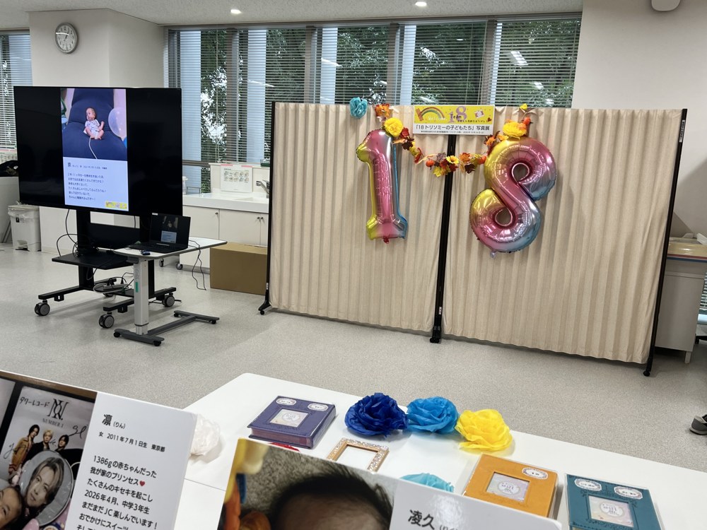 ベージュのパーテーションに「18」の虹色のバルーンと秋の葉の飾り、「18トリソミーの子どもたち写真展」の横断幕。左のモニターにはパネルの写真が映っている。手前の机には写真パネルと色紙のバラ