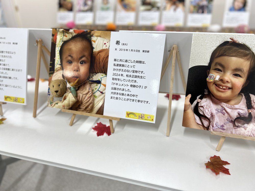 クマのぬいぐるみと一緒に写る赤ちゃんの写真パネル。「希(まれ)」の名前と、『ドキュメント 奇跡の子』が出版されたことを伝える家族の言葉が添えられている。となりには笑顔の女の子のパネル